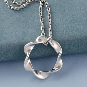 Peut inclure: Un collier argenté avec un pendentif en forme de cercle ouvert torsadé. Le pendentif est suspendu à une chaîne délicate. Le collier repose sur une surface bleu-gris, créant un contraste.