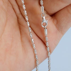 May include: A close-up of a silver-colored necklace with a barrel chain design. The chain is composed of small, textured, cylindrical beads linked together. A lobster clasp is visible at the end of the chain, ready for secure fastening.