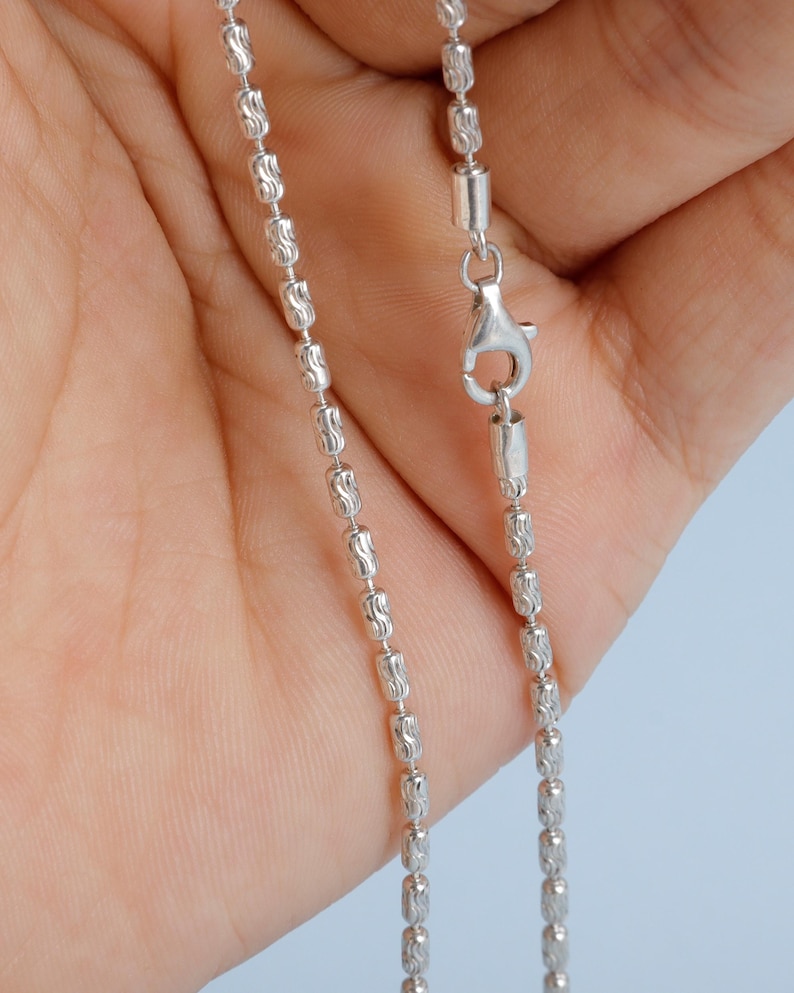 May include: A close-up of a silver-colored necklace with a barrel chain design. The chain is composed of small, textured, cylindrical beads linked together. A lobster clasp is visible at the end of the chain, ready for secure fastening.