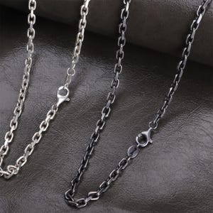 May include: Three silver-toned chain necklaces with lobster claw clasps. The chains have different finishes, including a bright silver and a darker, oxidized silver. The chains are displayed against a dark, textured background.