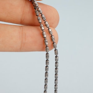 May include: Close-up of a silver necklace with a beaded design. The necklace features small, barrel-shaped beads strung together. The beads have a textured, etched pattern. The necklace is held against a light blue background.