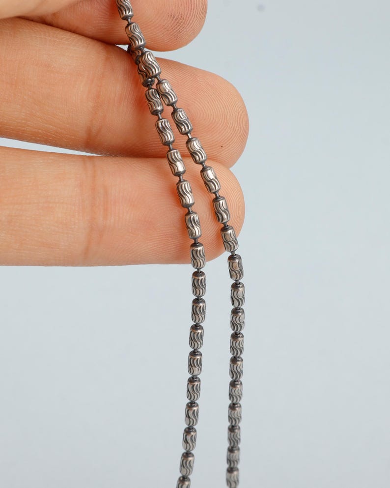 May include: Close-up of a silver necklace with a beaded design. The necklace features small, barrel-shaped beads strung together. The beads have a textured, etched pattern. The necklace is held against a light blue background.