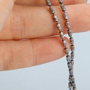 May include: Close-up of a silver necklace with a delicate chain. The chain is composed of small, cylindrical beads with a wavy pattern. The necklace is held against a light blue background.