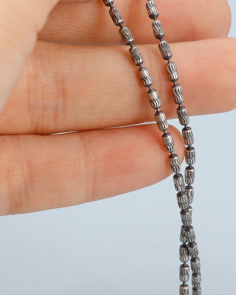 May include: Close-up of a silver necklace with a delicate chain. The chain is composed of small, cylindrical beads with a wavy pattern. The necklace is held against a light blue background.