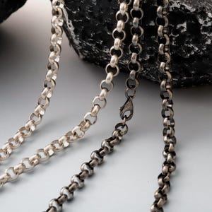 May include: Close-up of three silver-toned chain necklaces with a rounded link design. One chain has a clasp. The chains are displayed against a dark, textured background, highlighting the metallic shine of the jewelry.