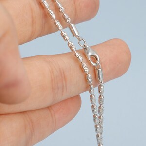 May include: A silver necklace with a delicate, beaded chain design. The chain is composed of small, textured beads and cylindrical links, and it has a secure clasp. The necklace is displayed against a light blue background.