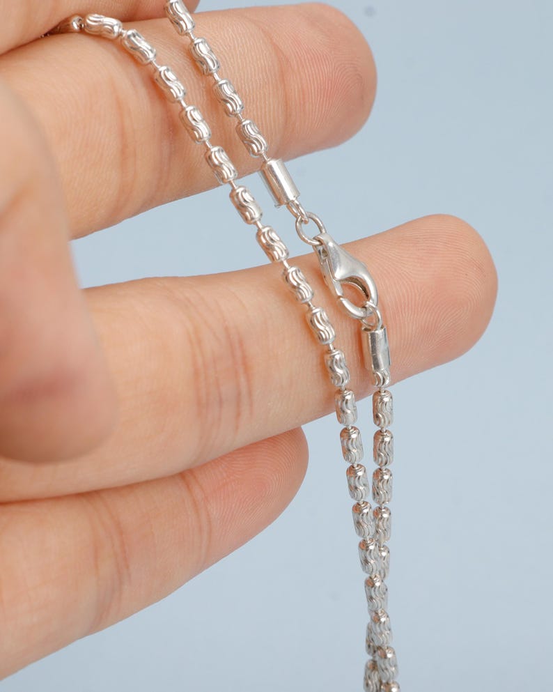 May include: A silver necklace with a delicate, beaded chain design. The chain is composed of small, textured beads and cylindrical links, and it has a secure clasp. The necklace is displayed against a light blue background.