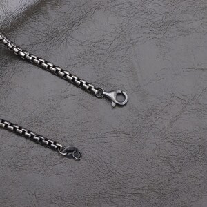 May include: A close-up of a silver-colored box chain necklace with a lobster clasp. The chain is made of small, square links and lies on a textured, dark gray surface. The necklace is a jewelry item.
