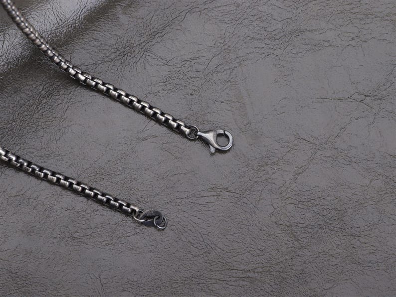 May include: A close-up of a silver-colored box chain necklace with a lobster clasp. The chain is made of small, square links and lies on a textured, dark gray surface. The necklace is a jewelry item.