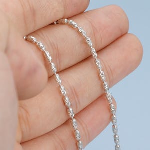 May include: A delicate silver necklace with a repeating pattern of small, textured beads. The necklace is held in a hand against a light blue background, showcasing its intricate design and reflective surface. The chain is approximately 18 inches long.