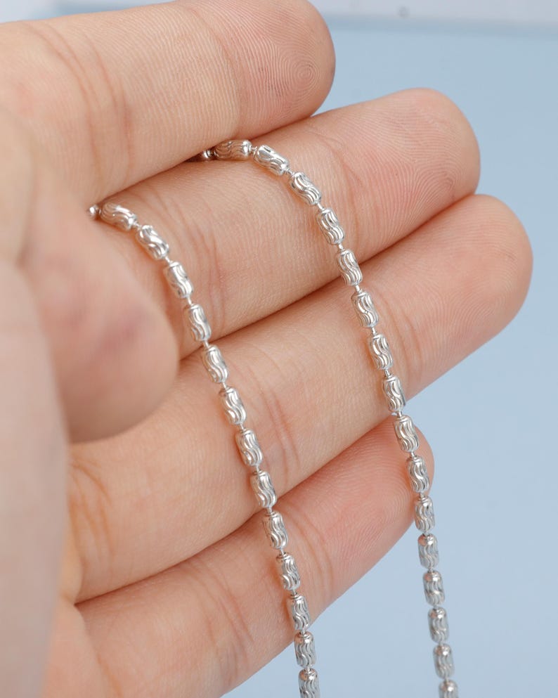 May include: A delicate silver necklace with a repeating pattern of small, textured beads. The necklace is held in a hand against a light blue background, showcasing its intricate design and reflective surface. The chain is approximately 18 inches long.