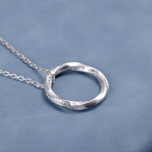 Puede incluir: Un collar de tono plateado con un colgante circular de diseño retorcido. La superficie del anillo de metal está grabada con números romanos, incluyendo VIII y IX, y está suspendido de una delicada cadena de eslabones de tono plateado.