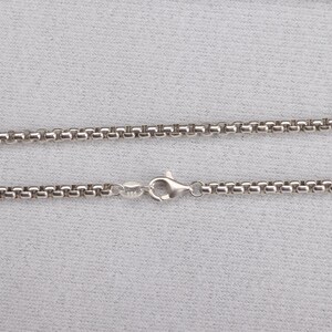 May include: A silver-colored necklace with a round box chain design. The chain is connected with a lobster clasp. The necklace is displayed on a light gray surface.