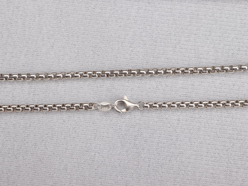May include: A silver-colored necklace with a round box chain design. The chain is connected with a lobster clasp. The necklace is displayed on a light gray surface.