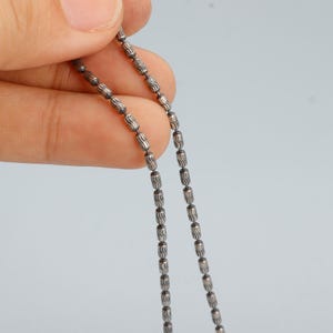 May include: Close-up of a silver-colored beaded chain. The chain is composed of small, barrel-shaped beads with etched designs. The chain is held against a light gray background, showcasing the intricate details of the jewelry.