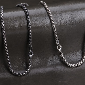 Puede incluir: Dos collares de cadena plateados con un diseño de eslabones cuadrados. Cada collar tiene un cierre de mosquetón. Las cadenas se muestran sobre una superficie oscura y texturizada.