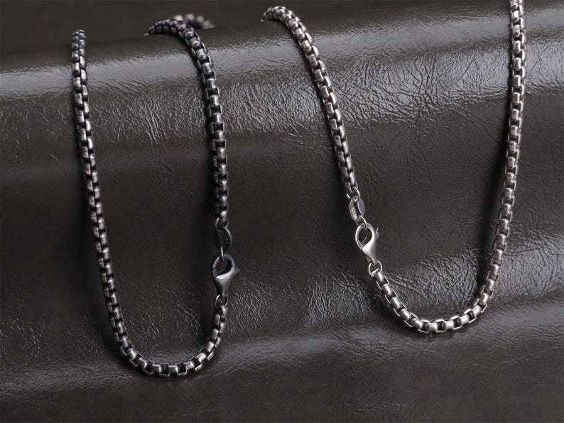 May include: Two silver-colored chain necklaces with a box link design. Each necklace has a lobster clasp closure. The chains are displayed on a dark, textured surface.