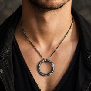 May include: A dark silver-toned metal pendant necklace featuring a circular ring charm with a twisted, Mobius-like design and intricate geometric Greek key patterns engraved along the surface, suspended from a matching dark silver-toned chain.