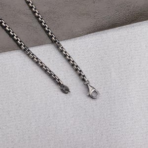 May include: A silver-toned necklace with a box chain design. The necklace features a lobster clasp closure and a small tag at the end of the chain. The chain is displayed on a textured white surface.
