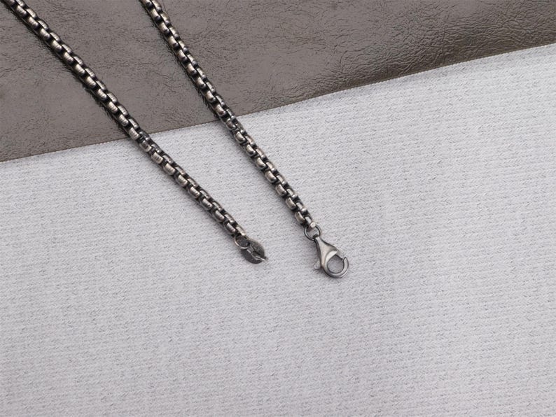 May include: A silver-toned necklace with a box chain design. The necklace features a lobster clasp closure and a small tag at the end of the chain. The chain is displayed on a textured white surface.