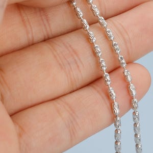 May include: Close-up of a silver chain with a repeating pattern of elongated beads. The chain is held against a light blue background, showcasing its intricate design and reflective surface. The chain's links are detailed and evenly spaced.