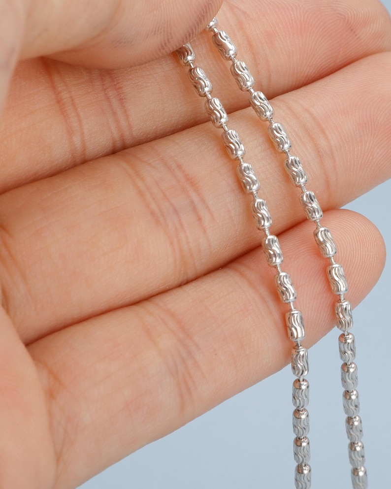 May include: Close-up of a silver chain with a repeating pattern of elongated beads. The chain is held against a light blue background, showcasing its intricate design and reflective surface. The chain's links are detailed and evenly spaced.