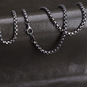 May include: A silver-toned box chain necklace with a lobster clasp. The chain is composed of small, square links and rests on a dark, textured surface. The necklace is a jewelry item.