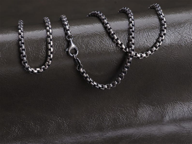 May include: A silver-toned box chain necklace with a lobster clasp. The chain is composed of small, square links and rests on a dark, textured surface. The necklace is a jewelry item.
