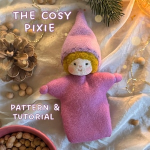 Könnte beinhalten: Eine handgefertigte Feenpuppe aus Filz in Rosatönen, mit einer spitzen Mütze und gelbem Haar. Die Puppe hat ein süßes, einfaches Gesicht. Der Text "THE COSY PIXIE" und "PATTERN & TUTORIAL" sind zu sehen. Das Bild hat eine warme, einladende Ästhetik.