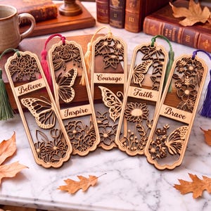 Op de afbeelding: Vijf lasergesneden houten bladwijzers met gedetailleerde bloem- en vlinderontwerpen. Elke bladwijzer toont een uniek woord: Believe, Inspire, Courage, Faith en Brave. Ze zijn afgewerkt met kleurrijke stoffen kwastjes in groen, roze, geel en paars.