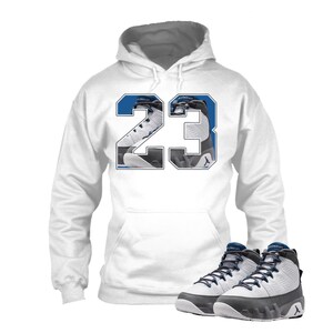 May include: White hoodie with the number 23 in blue and black, featuring a graphic of a basketball shoe. A pair of black, white, and blue basketball shoes are in the foreground. The shoes have a high-top design.