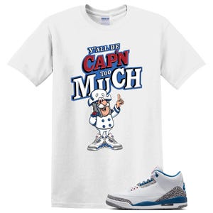 May include: A white short-sleeve t-shirt featuring the text Y'ALL BE CAP'N TOO MUCH in blue and red lettering above a cartoon character wearing a white uniform and blue and grey sneakers, accompanied by a matching white, blue, and grey sneaker.