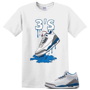 May include: A white short-sleeve t-shirt featuring a graphic of a white, blue, and grey sneaker with a dripping effect. Above the sneaker, the text 3'S is written in a blue dripping font. A matching sneaker is positioned at the bottom right.
