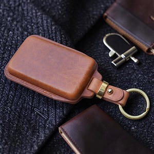 Op de afbeelding: Een rechthoekig bruin lederen sleuteletui met een gladde afwerking en zichtbare stiksels langs de randen. Het etui is voorzien van een goudkleurige metalen connector en een ronde goudkleurige sleutelring, bevestigd aan een bijpassende leren riem.