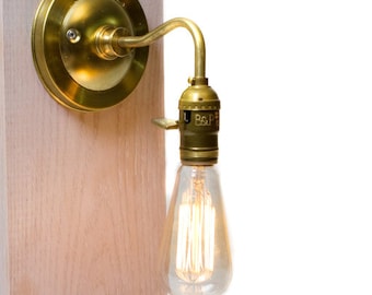 Könnte beinhalten: Eine Wandleuchte aus Messing mit einer klaren Glühbirne aus Glas. Die Leuchte hat einen runden Sockel und einen gebogenen Arm. Die Glühbirne ist eine Glühbirne mit Glühfaden im Vintage-Stil.