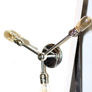 May include: A silver wall sconce with three light bulbs. The sconce has a circular base and three arms that extend out to hold the bulbs. The bulbs are all turned on and are emitting a warm, yellow light.
