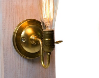 Könnte beinhalten: Eine goldene Wandleuchte mit einer klaren Glühbirne. Die Leuchte hat einen runden Sockel und einen gebogenen Arm.