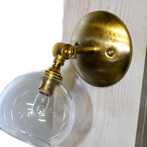 Puede incluir: Una lámpara de pared dorada con una pantalla de vidrio transparente y una bombilla en su interior. La lámpara está montada en una pared blanca.