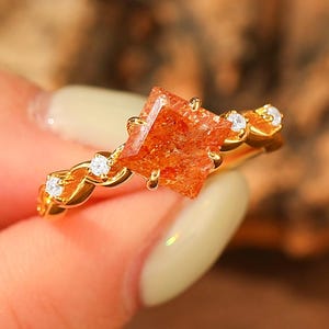 May include: A gold ring featuring a square-cut, orange gemstone as the centerpiece. The band is adorned with small, clear gemstones and a decorative, twisted design. The ring is held by a person.