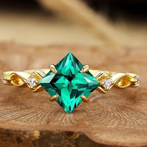 May include: A gold ring featuring a square-cut, vibrant green gemstone as the centerpiece. The band has a delicate, interwoven design with small, sparkling accent stones. The ring is set against a natural wood surface.