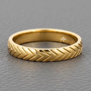 May include: A gold ring featuring a continuous, repeating chevron or herringbone pattern engraved around the entire outer band. The metal has a polished, reflective finish, and the interior of the band displays the small, clear text ANAGRA.
