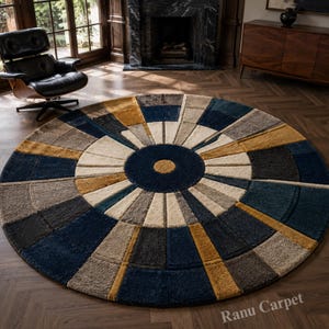 May include: A circular area rug featuring a geometric sunburst pattern in shades of navy blue, mustard yellow, cream, and charcoal gray. The design consists of concentric rings and radiating segments with a textured pile. The text Ranu Carpet is visible.