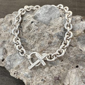 May include: A silver-colored chain bracelet with a cross-shaped clasp. The bracelet features a series of interconnected, rounded links. The cross clasp has the word "Custom" engraved on it. The bracelet is displayed on a gray, textured surface.