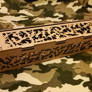 Può includere: Una scatola rettangolare in legno con intricati disegni tagliati al laser sul coperchio e sui lati. La scatola è realizzata in legno marrone chiaro e poggia su uno sfondo mimetico. I motivi ritagliati creano un effetto decorativo.