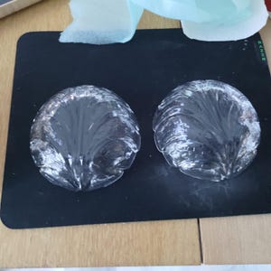 May include: Two clear, glass, shell-shaped objects with a textured, leaf-like design. The objects are on a black surface, with a hint of a wooden surface in the background. The glass pieces are reflective and appear to be decorative.