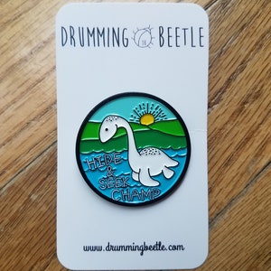 May include: A white enamel pin with a green, blue, and yellow illustration of a Loch Ness Monster with the text "Hide & Seek Champ" and the website "www.drummingbeetle.com".