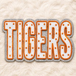 Könnte beinhalten: Ein großer, rechteckiger Aufnäher mit dem Wort TIGERS in fetten Blockbuchstaben. Die Buchstaben sind weiß mit einer schimmernden Paillettentextur, mit leuchtend orangefarbenem Garn umrandet und mit kleinen, orangefarbenen, sternförmigen Stickereien versehen.