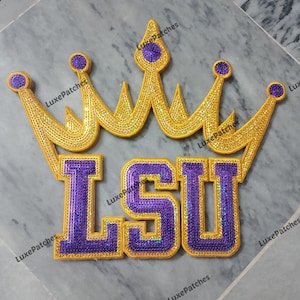 May include: A decorative patch featuring the letters LSU in purple sequins with a gold sequin border, positioned beneath a large, five-pointed crown design also crafted from gold and purple sequins. The patch has a distinct, layered aesthetic.
