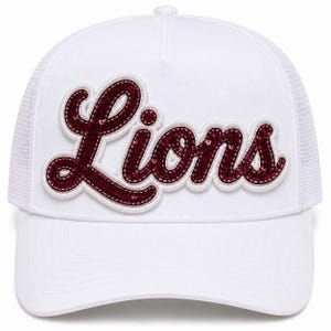 Puede incluir: Una gorra tipo trucker blanca con una aplicación prominente en letra cursiva en el panel frontal. La palabra "Lions" está bordada en granate oscuro, adornada con lentejuelas a juego y delineada con un borde grueso de tela blanca en relieve.