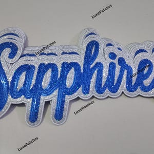 May include: A layered script patch featuring the word Sapphires in bright blue sequins with a white glitter border. The design has a dimensional appearance with multiple stacked layers of white and blue material, creating a thick, raised effect.